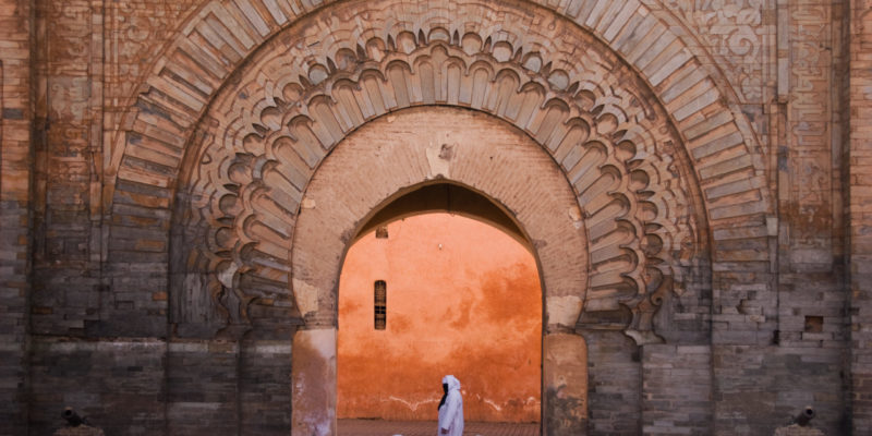 Woman in traditional arab clothing with veil passing by the Bab Anaou gate in Marrakech. The woman walks right in the middle of the gate that is colored orange by the late sunlight.